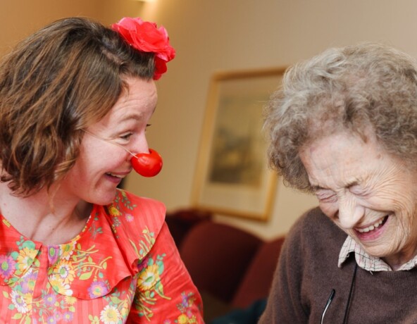 Woman dressed as a clown making an older person laugh.