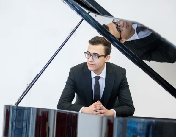 Young man in suit and tie standing behind a black grand piano.